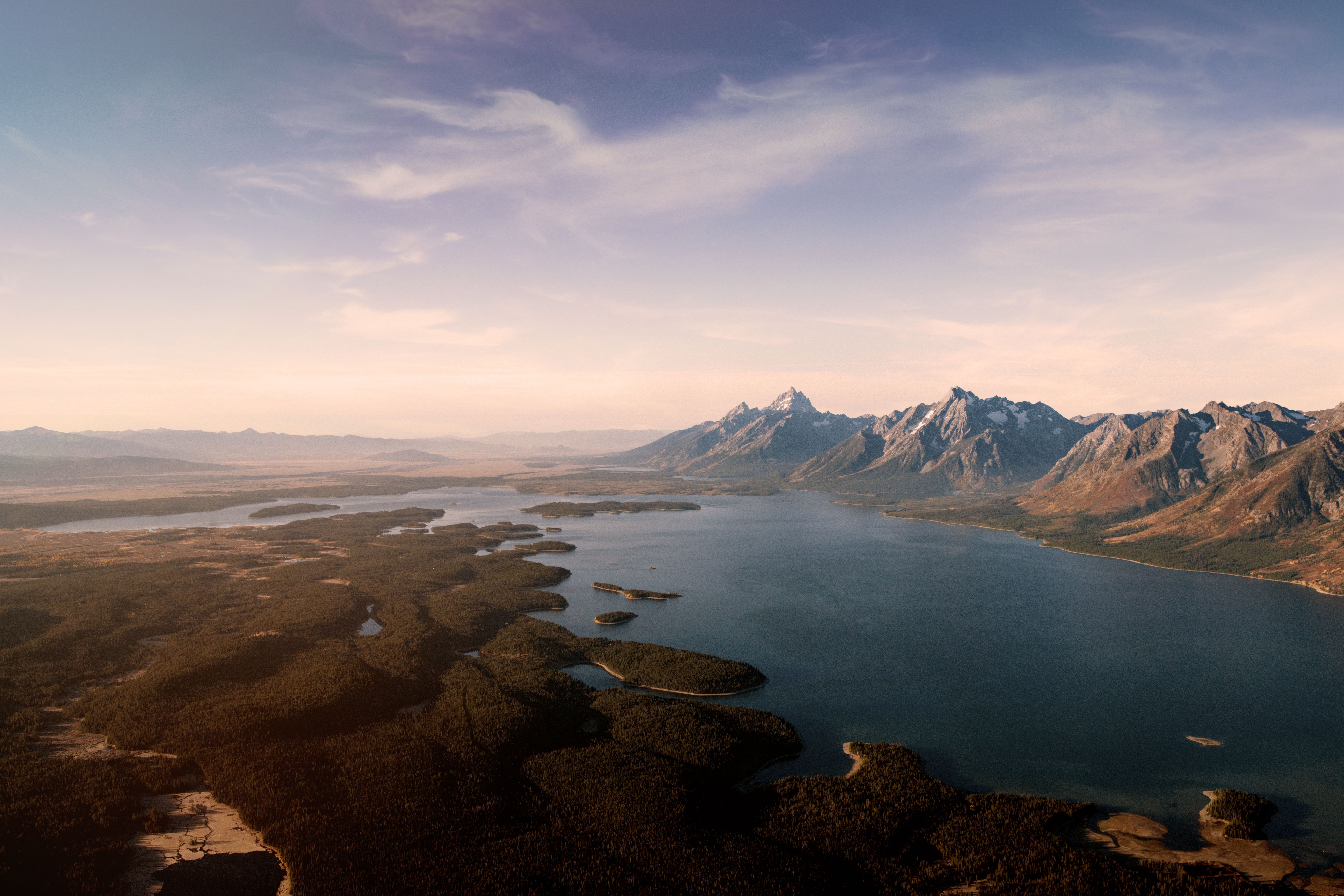 Aerial view of the Teton range above Jackson Lake at golden hour, with the forested valley below