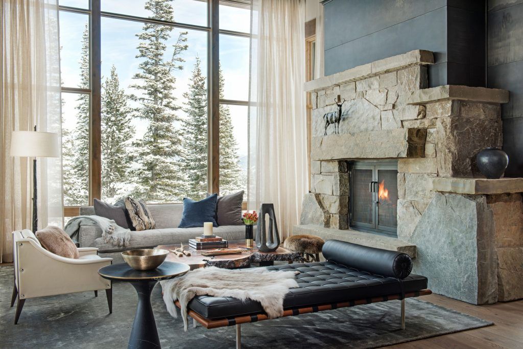 A mountain home living room with a lit stone fireplace and floor-to-ceiling windows framing snowy pines